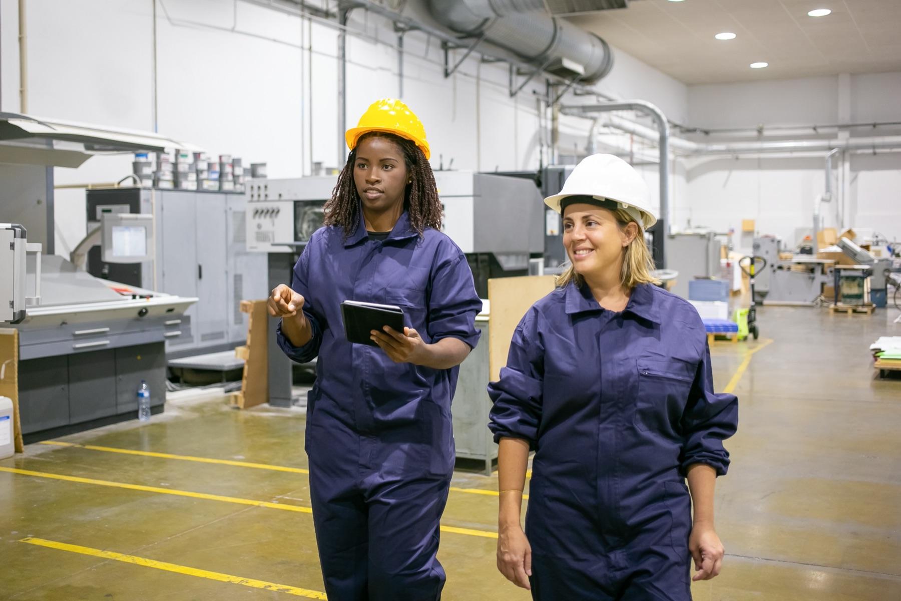 Two factory workers in coveralls and hard hats walking through the shop floor, one holding a tablet